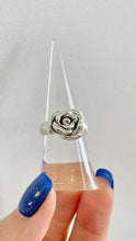 Load image into Gallery viewer, A silver ring shaped like a rose, held on a clear cone display stand by a hand with glittery blue nail polish against a light gray textured background.

